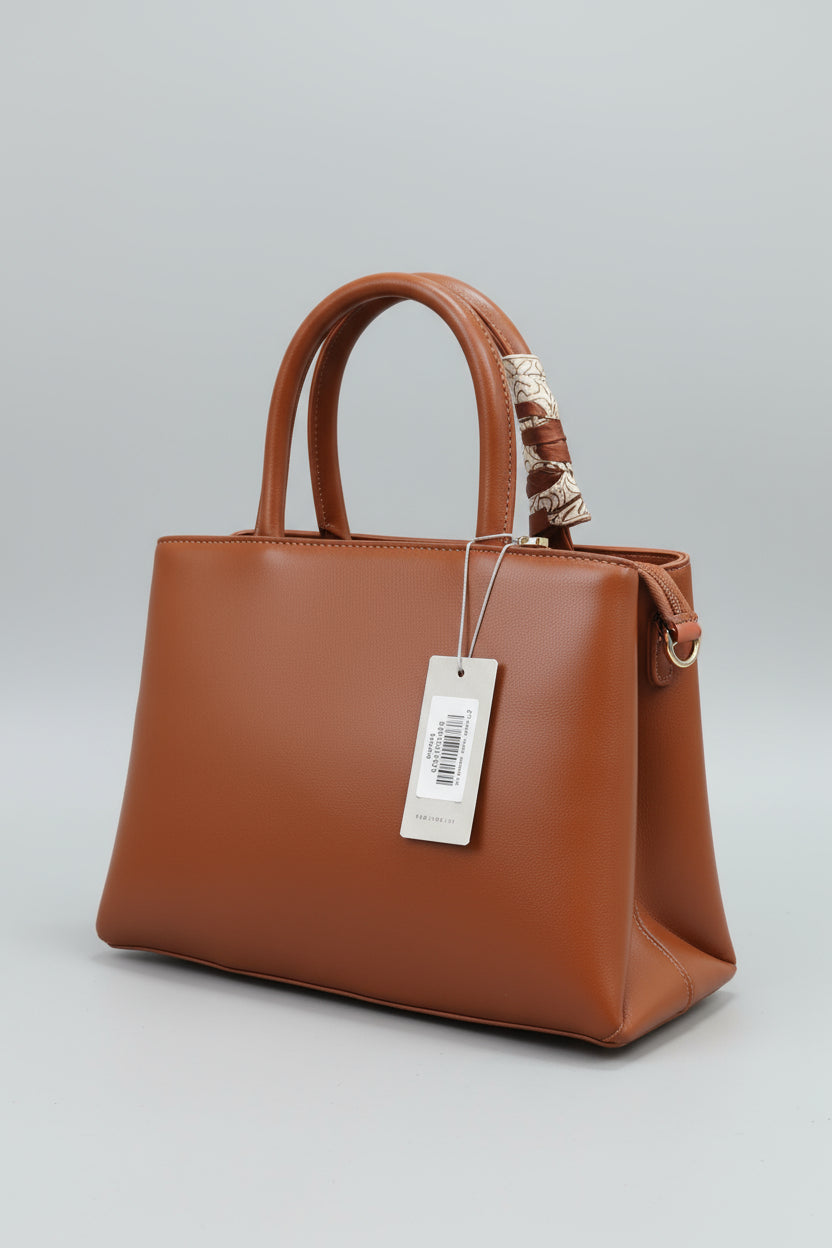 Brown handbag held by a hand with a visible brand tag against a green grass background.